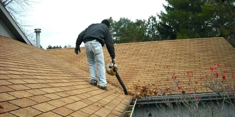 Worker clearing leaves and debris from the roof valley and gutter line of a shingle roof